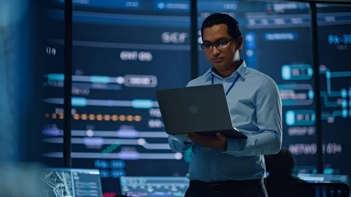 Cybersecurity analyst monitoring data on a laptop in a security operations center.
